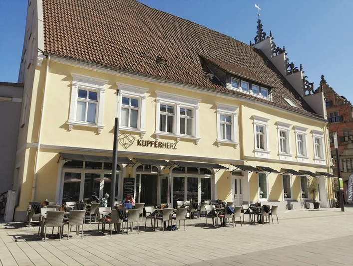 Ein historisches Gebäude mit gelber Fassade beherbergt ein Café mit Terrasse und weißen Sitzmöbeln.