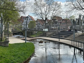 Hängebrücke am Wasserspielplatz über einem Bach vor Fachwerkhäusern und Bäumen