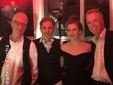 Vier Personen lächeln in die Kamera, sie tragen elegante Kleidung bei einem stimmungsvoll beleuchteten Event.