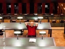 Eine Person sitzt entspannt an einem großen Tisch in einer modernen, gut beleuchteten Bibliothek.