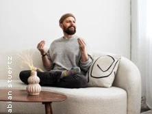 Ein Mann mit Bart sitzt entspannt auf einem hellen Sofa, umgeben von einem minimalistischen Raum.