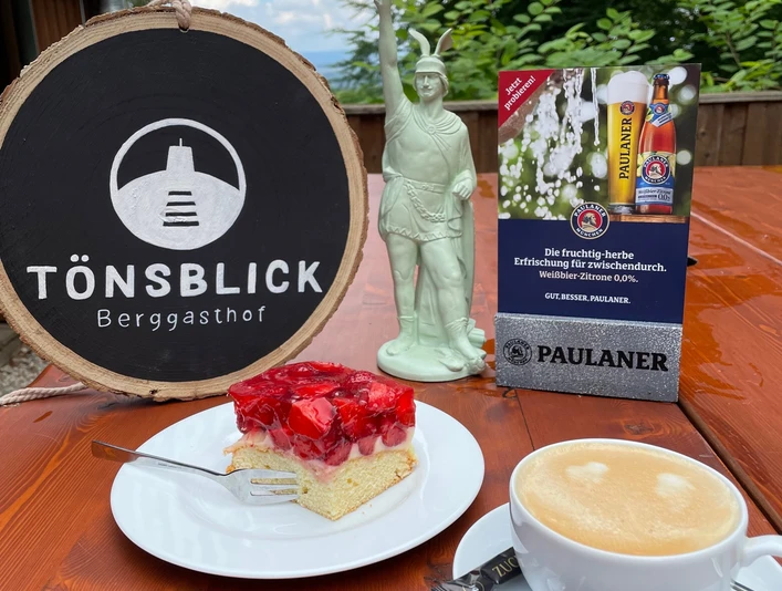 Berggasthof Tönsblick Vorne ein Stück Erdbeerkuchen und Kaffee auf einem Tisch, hinten ein Schild "Tönsblick".