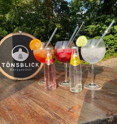 Drei Gläser mit Cocktails sowie zwei Flaschen Getränk auf Holztisch im Freien beim TönsBlick.