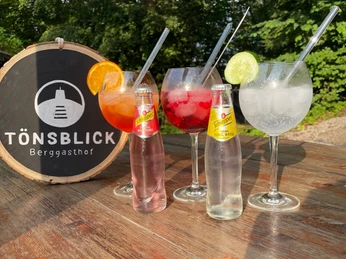 IMG_2601.jpg Drei Gläser mit Cocktails sowie zwei Flaschen Getränk auf Holztisch im Freien beim TönsBlick.