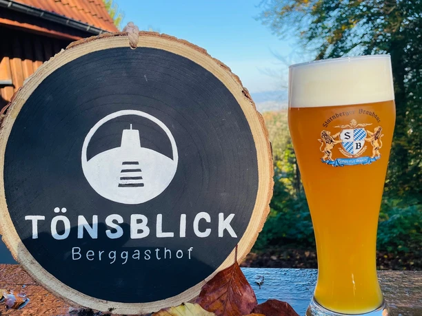 IMG_3756.jpg Ein Weizenglas, gefüllt mit Schaumkrone, steht neben einem Holzschild des Berggasthofs Tönsblick.