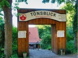 IMG_9694.jpg Eingang des Berggasthofs TönsBlick mit hölzernem Rundbogen, umgeben von grünem Laub und Bäumen.