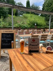 Herzlichen Willkommen im Berggasthof Tönsblick Biergarten mit rustikalen Holztischen, Paulaner Bier, Speisekarte und umgeben von grüner Natur.