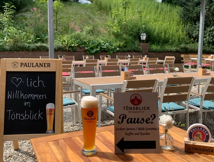 Herzlichen Willkommen im Berggasthof Tönsblick Biergarten mit rustikalen Holztischen, Paulaner Bier, Speisekarte und umgeben von grüner Natur.