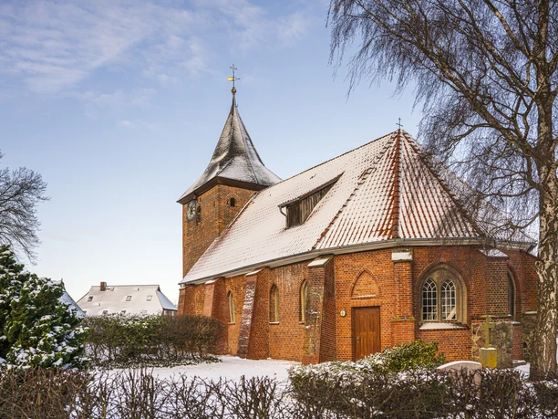 Historic red-brick church with snow-covered roof and tower, surrounded by trees and winter landscape.
