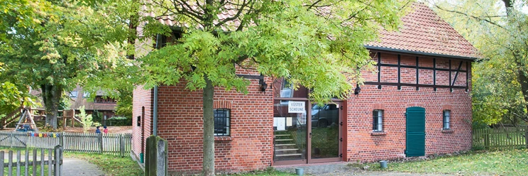 kuesterscheune_schmal.jpg Das Bild zeigt ein traditionelles Backsteingebäude, die Küsterscheune, umgeben von Bäumen im Garten.