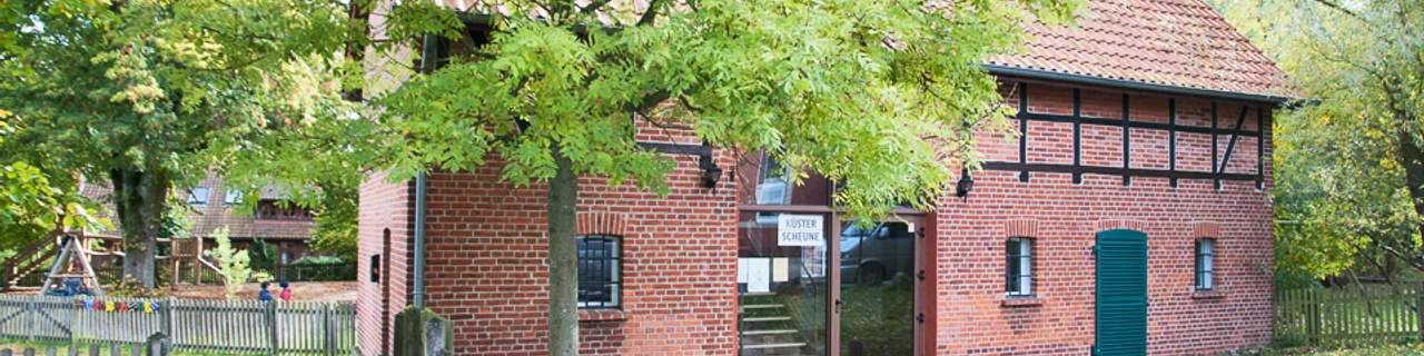 kuesterscheune_schmal.jpg Das Bild zeigt ein traditionelles Backsteingebäude, die Küsterscheune, umgeben von Bäumen im Garten.