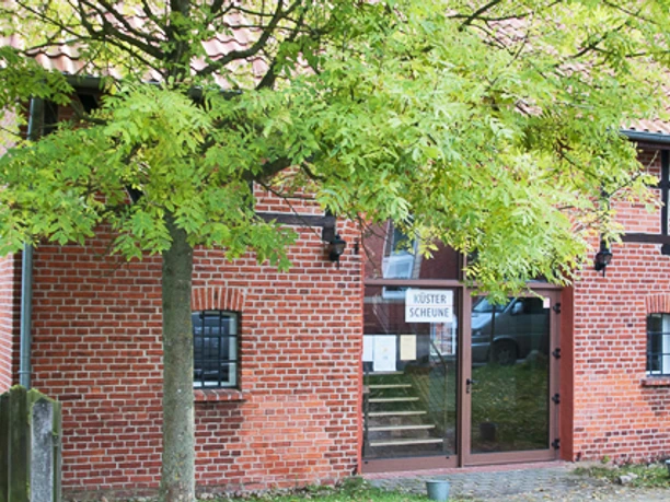 kuesterscheune_schmal.jpg Das Bild zeigt ein traditionelles Backsteingebäude, die Küsterscheune, umgeben von Bäumen im Garten.