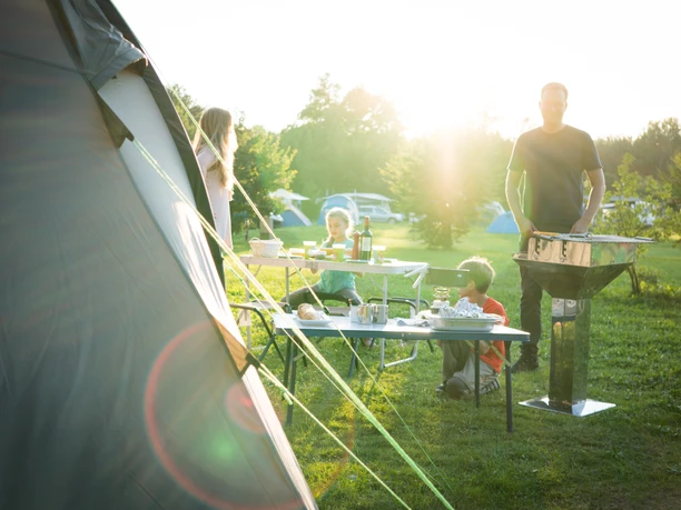 Camping Familie beim Campen, genießt Mahlzeit im Freien, Sonnenuntergang im Hintergrund, Zelt rechts.