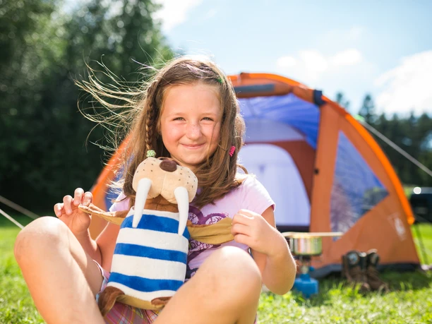 Camping Ein lachendes Mädchen sitzt vor einem orangefarbenen Zelt, hält ein Plüschtier.