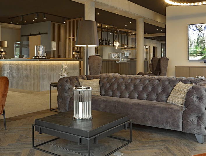 Hotel-Zeitgeist_Warburg_neu.JPG Moderner Hotel-Lounge-Bereich mit grauen gepolsterten Sofas, rustikalen Holzmöbeln und gedämpfter Beleuchtung.