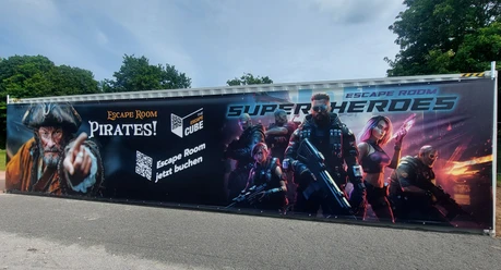 Container mit Werbeaufdruck für Piraten- und Superhelden-Escape Rooms, umgeben von Bäumen.