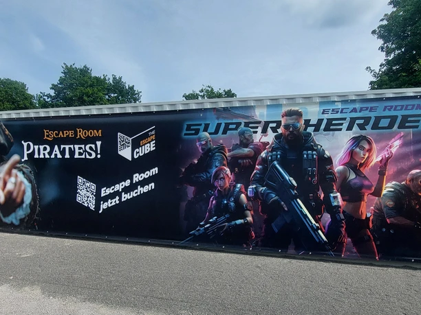 Escape Cube SchiederSee.jpg Container mit Werbeaufdruck für Piraten- und Superhelden-Escape Rooms, umgeben von Bäumen.