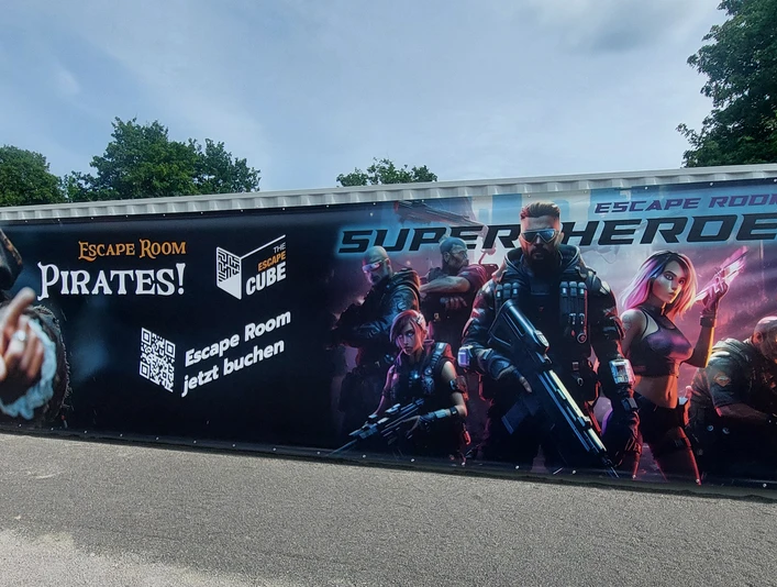 Escape Cube SchiederSee.jpg Container mit Werbeaufdruck für Piraten- und Superhelden-Escape Rooms, umgeben von Bäumen.