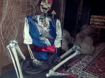Skelett in Piratenkleidung sitzt auf Teppich, neben Kisten und Netzen in einem Escape Room.