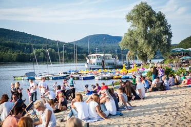 Strandbesucher am Schiedersee entspannen, während Segelboote und ein Ausflugsdampfer vorbeiziehen.