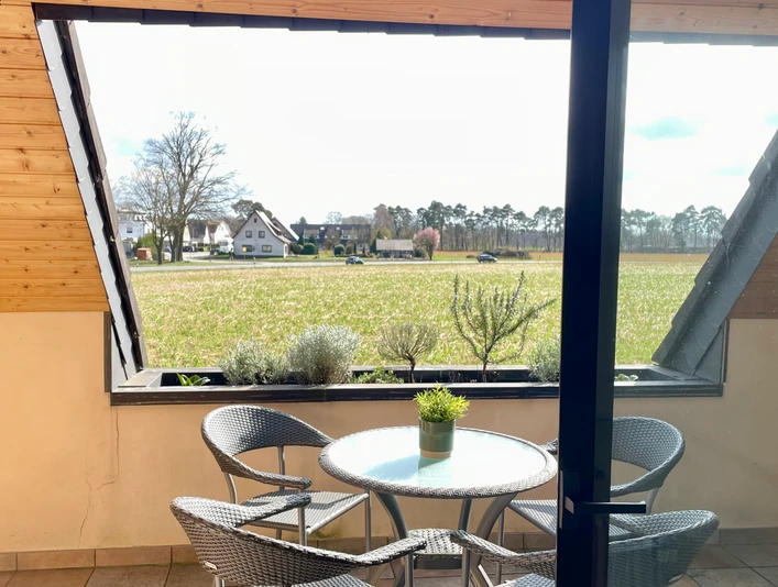 Fewo_Liemke_Balkon_SHSCC-BY-SA_Teutoburger_Wald_Stadt_Schloss_Holte-Stukenbrock Ein Balkon mit Tisch und vier Stühlen bietet Blick auf ein weites Feld unter einem bewölkten Himmel.