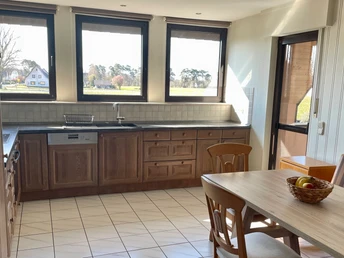 Helle Wohnküche mit Holzmöbeln, drei Fenstern, Fliesenboden und einem einladenden Esstisch.