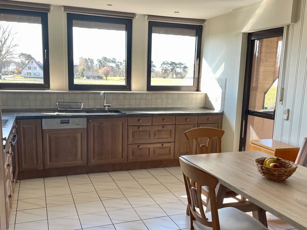 Fewo_Liemke_Wohnküche_SHSCC-BY-SA_Teutoburger_Wald_Stadt_Schloss_Holte-Stukenbrock Helle Wohnküche mit Holzmöbeln, drei Fenstern, Fliesenboden und einem einladenden Esstisch.