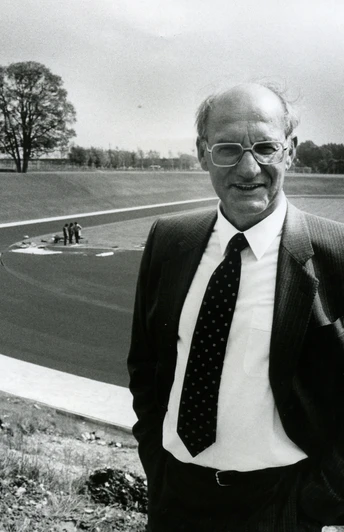 Eine ältere Person im Anzug steht vor einem leeren Sportplatz mit einer umlaufenden Laufbahn.