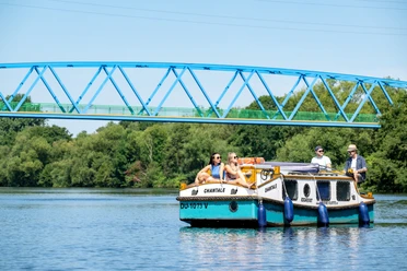 Ein kleines Ausflugsboot auf einem ruhigen Fluss mit grünen Bäumen im Hintergrund und einer blauen Brücke darüber. Mehrere Personen sitzen entspannt an Bord, während das Boot sanft im Wasser gleitet.