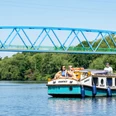 Escargot-Tour, Mülheim an der Ruhr Ein kleines Ausflugsboot auf einem ruhigen Fluss mit grünen Bäumen im Hintergrund und einer blauen Brücke darüber. Mehrere Personen sitzen entspannt an Bord, während das Boot sanft im Wasser gleitet.