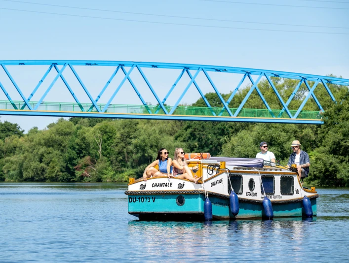Escargot-Tour, Mülheim an der Ruhr Ein kleines Ausflugsboot auf einem ruhigen Fluss mit grünen Bäumen im Hintergrund und einer blauen Brücke darüber. Mehrere Personen sitzen entspannt an Bord, während das Boot sanft im Wasser gleitet.