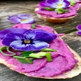 Waldkräuterey Ein Baguette mit einer leuchtend violetten Aufstrichcreme, garniert mit essbaren Blüten und Schnittlauch.