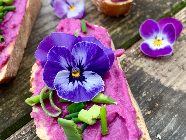 Waldkräuterey Ein Baguette mit einer leuchtend violetten Aufstrichcreme, garniert mit essbaren Blüten und Schnittlauch.