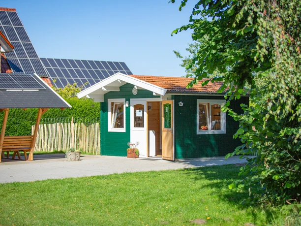 In't Euwer Grünes Holzhaus mit rotem Satteldach, offenem Eingang und umgeben von Bäumen und Sonnenschein.