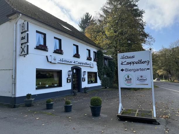 Haus Koppelberg Außenansicht 2 Weißes zweistöckiges Gebäude mit Biergarten in ländlicher Umgebung und blauem Himmel.