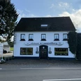 Haus Koppelberg Außenansicht Gasthaus mit weißen Wänden, dunklem Dach und grüner Rankenverzierung, daneben eine Straße.