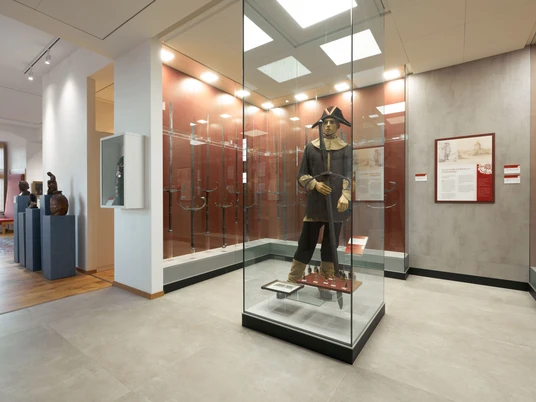 11053_Burgmuseum_0010.jpg Ein Museumsraum mit Glasvitrinen, enthält historische Rüstungen und Waffen, rot und weiß gestaltet.