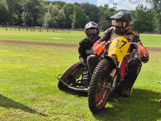 Grasbahnrennen Fahrer.jpg Zwei Motorsportler fahren mit einem Motorradgespann über eine Grasbahn, umgeben von Wald.