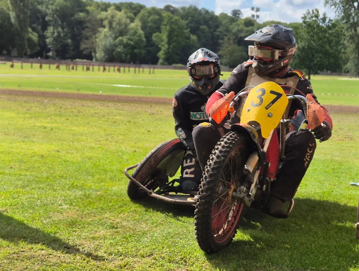 Grasbahnrennen Fahrer.jpg Zwei Motorsportler fahren mit einem Motorradgespann über eine Grasbahn, umgeben von Wald.