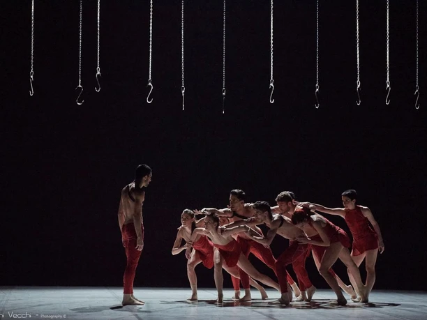 Schrit_tmacher Festival Aachen Eine Gruppe Tänzer in roten Kostümen formt eine dynamische Formation, während ein Solotänzer am linken Bildrand steht.