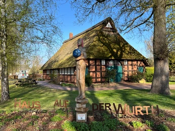 Weltenträgerin Bronzeskulptur einer stehenden Frau mit Weltkugel vor traditionellem Reetdachhaus im Grünen.