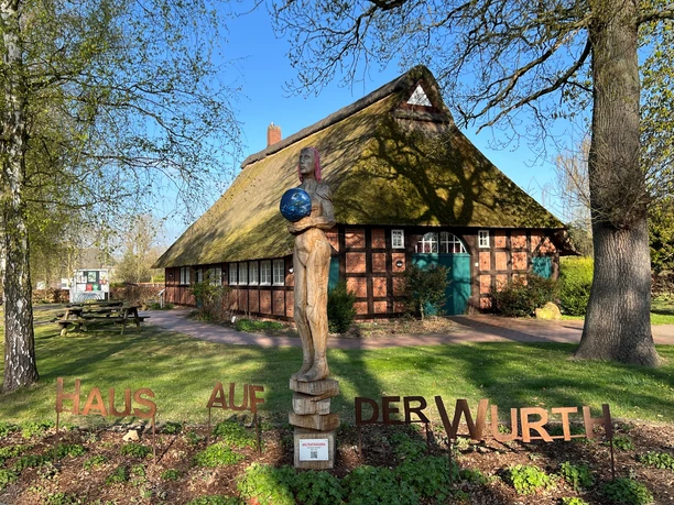 Weltenträgerin Bronzeskulptur einer stehenden Frau mit Weltkugel vor traditionellem Reetdachhaus im Grünen.