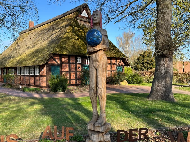 Weltenträgerin Eine hölzerne Skulptur eines Menschen, der eine blaue Erdkugel hält, vor einem Fachwerkhaus.