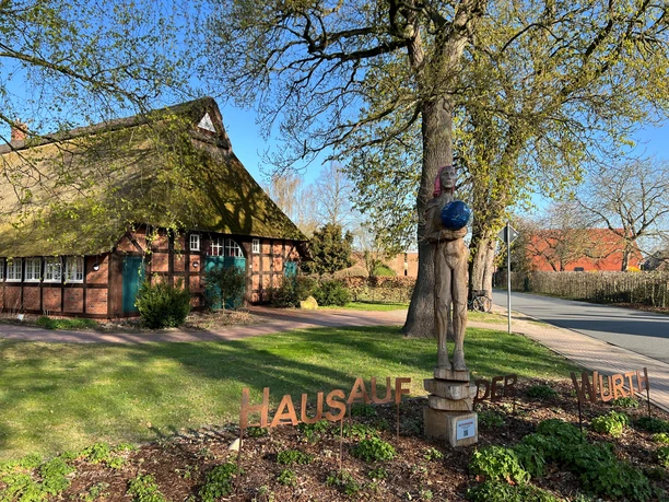 Weltenträgerin Holzskulptur einer Frau, die eine blaue Kugel hochhält, vor einem alten Fachwerkhaus.