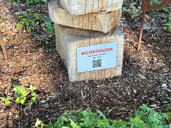 Weltenträgerin Eine Holzskulptur mit der Aufschrift "Weltenträgerin" von Uwe Oswald, umgeben von Grünpflanzen.