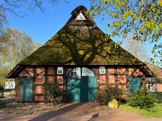Haus auf der Wurth Fachwerkhaus mit Reetdach und grüner Tür, umgeben von Bäumen, in frühlingshafter Landschaft.
