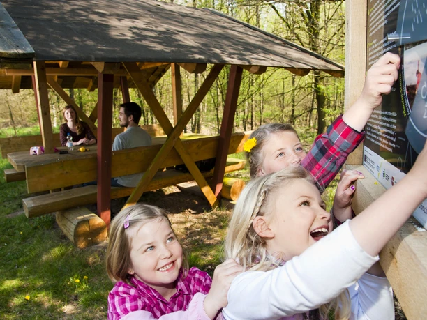 Walderlebnispfad Fullener Wald bei Meppen ©Naturpark Moor-Veenland (2).jpg Kinder erkunden eine Mitmachtafel im Wald, während Erwachsene an einem Holztisch im Hintergrund sitzen.
