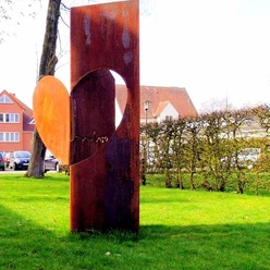 haller_herz_skulptur.jpg Eine rostige Skulptur mit Herz-Motiv steht auf grasbewachsenem Platz vor einer Hecke, Häuser im Hintergrund.