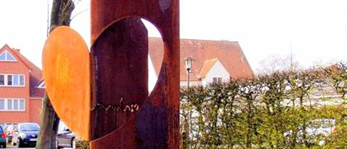 haller_herz_skulptur.jpg Eine rostige Skulptur mit Herz-Motiv steht auf grasbewachsenem Platz vor einer Hecke, Häuser im Hintergrund.