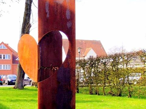 haller_herz_skulptur.jpg Eine rostige Skulptur mit Herz-Motiv steht auf grasbewachsenem Platz vor einer Hecke, Häuser im Hintergrund.
